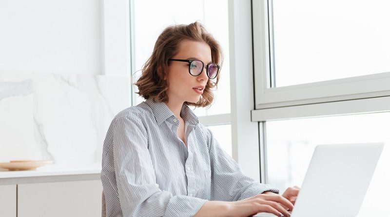 Photo of young concentrated woman in striped shirt using laptop while siting at table in light apartment