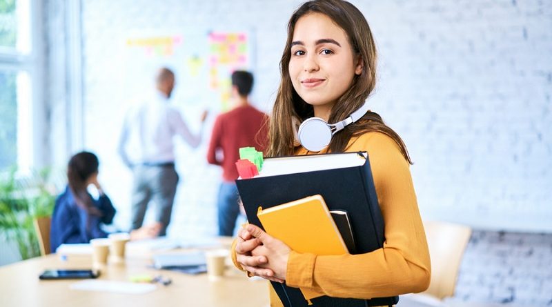 Female student standing in classroom holding books. Portrait of young woman looking at camera indoors