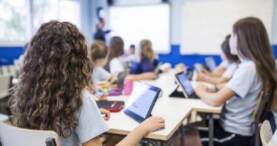 girl with brown curly hair studies in class with her tablet next to her classmates while they listen to the teacher