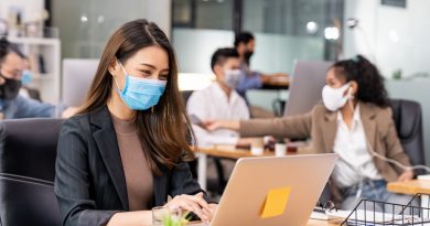 Portrait of asian office employee businesswoman wear protective face mask work in new normal office with interracial team in background as social distance practice prevent coronavirus COVID-19.