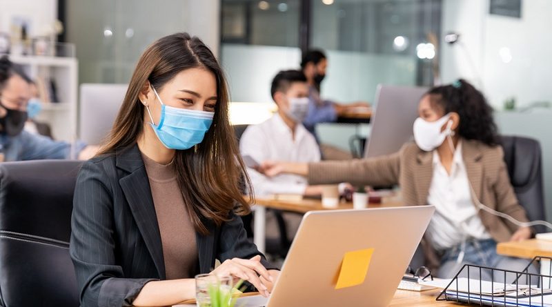 Portrait of asian office employee businesswoman wear protective face mask work in new normal office with interracial team in background as social distance practice prevent coronavirus COVID-19.