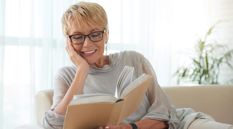 Senior blond woman in glasses resting on sofa and reading a book