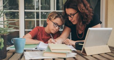 Young student doing homework at home with school books, newspaper and digital pad helped by his mother. Mum writing on the copybook teaching his son. Education, family lifestyle, homeschooling concept