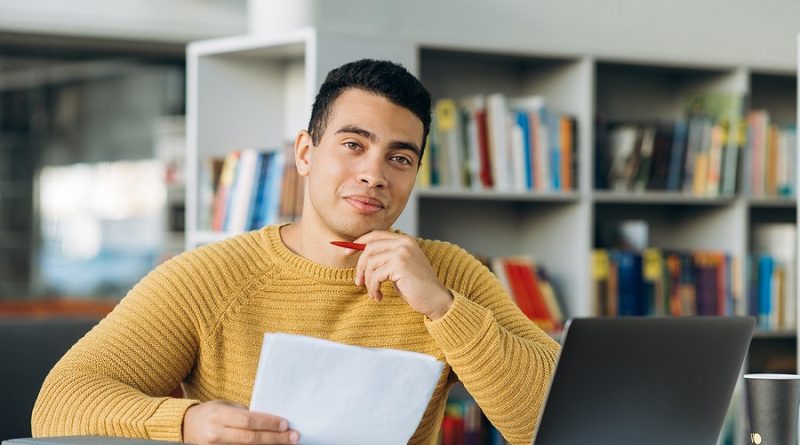 Portrait of young adult hispanic male freelancer or student is looking at the camera with smile. Attractive business man sits at the work desk, making notes, learning online
