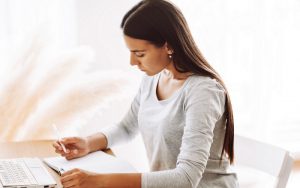 A beautiful student girl is sitting at a desk and making notes in a notebook, using a laptop to prepare for classes. Modern technologies, online education, training.