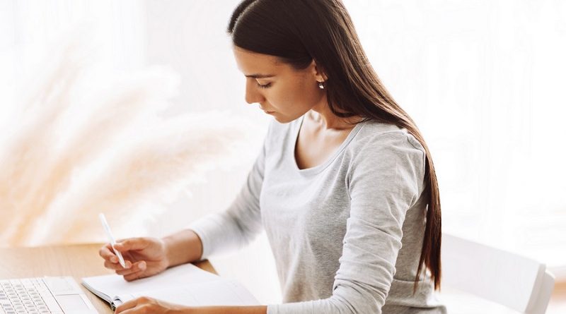 A beautiful student girl is sitting at a desk and making notes in a notebook, using a laptop to prepare for classes. Modern technologies, online education, training.