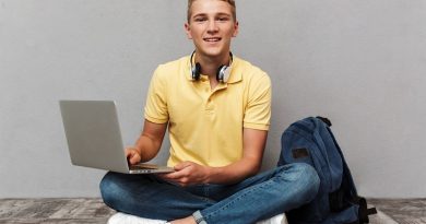 Portrait of a smiling casual teenage boy with backpack using laptop computer while sitting isolated over gray background