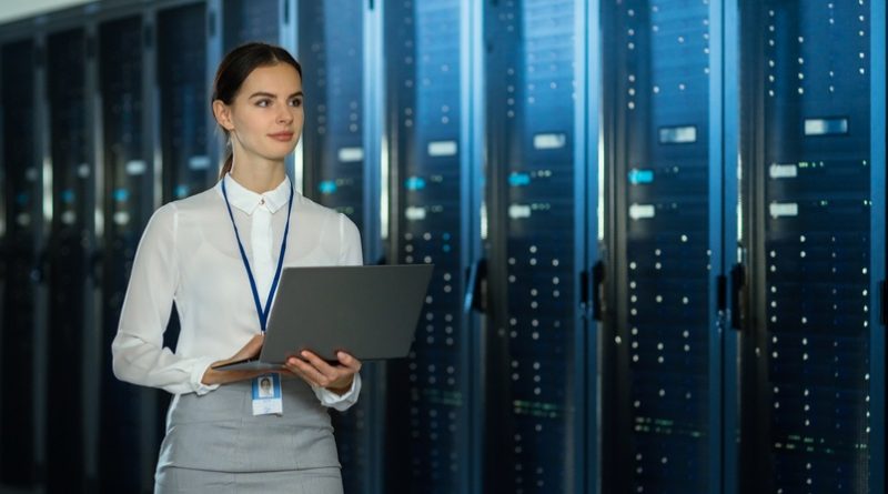 Beautiful Data Center Female IT Technician Walking Through Server Rack Corridor with a Laptop Computer. She is Visually Inspecting Working Server Cabinets.