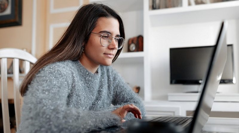 Young cheerful woman wearing a sweater in her home is sitting in her living room while browsing in the internet in the laptop.