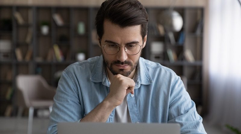 Thoughtful young male manager in eyeglasses looking at laptop screen, considering problem solution, preparing research report, analyzing sales statistic, developing strategy, working on online project
